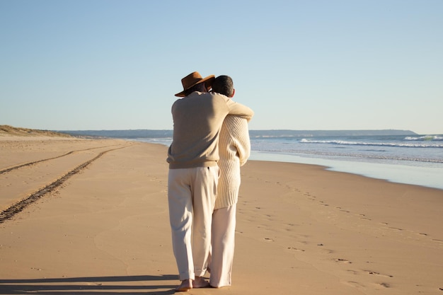 Senior paar wandelen aan de kust op warme herfstmiddag. Achteraanzicht van man in cowboyhoed knuffelen kortharige vrouw terwijl verliefde paar tijd doorbrengen op het strand. Liefde, saamhorigheid concept