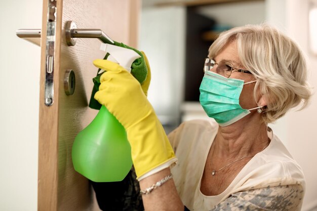 Senior vrouw desinfecteert deurklink tijdens coronavirus pandemie
