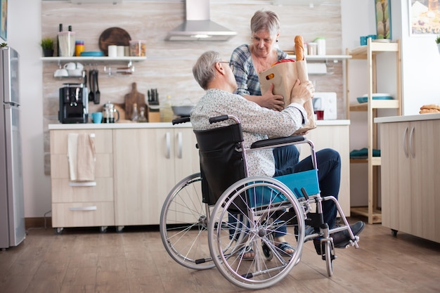 Senior vrouw die boodschappentas neemt van gehandicapte man in rolstoel. Volwassen mensen met verse groenten van de markt. Leven met een gehandicapte met een loophandicap