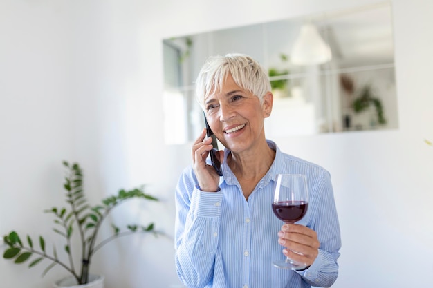 Senior vrouw die een glas wijn drinkt terwijl ze haar telefoon gebruikt Gelukkige vrouw die thuis ontspant, drinkt een glas wijn en praat op haar smartphone