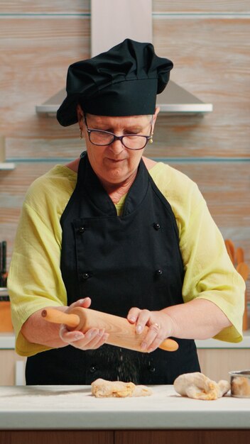 Senior vrouw die het deeg uitrolt en zelfgebakken brood bereidt. Gelukkige bejaarde chef-kok met bonete die rauwe ingrediënten bereidt voor het bakken van traditionele pizzabestrooiing, zeven van bloem op tafel in de keuken.