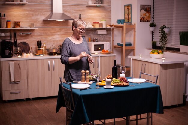 Senior vrouw die kaarsen aansteekt in de keuken voor een romantisch diner met man. Oudere vrouw die haar man wacht op een romantisch diner. Rijpe vrouw die feestelijke maaltijd voorbereidt voor jubileumviering.