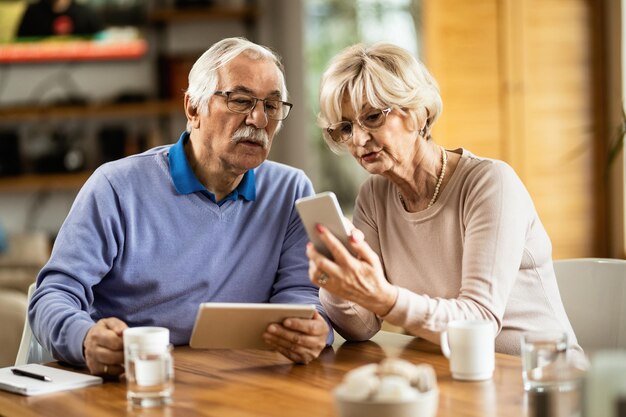 Senior vrouw die mobiele telefoon gebruikt en sms-bericht laat zien aan haar man die thuis digitale tablet gebruikt