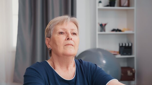Senior vrouw doet ademhalingsoefening tijdens het mediteren in de woonkamer. Gepensioneerde bejaarde oefent thuis sportactiviteit uit op de pensioengerechtigde leeftijd