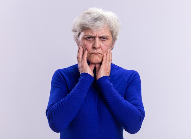 Senior vrouw in blauwe coltrui die naar de camera kijkt met een fronsend gezicht op een witte achtergrond