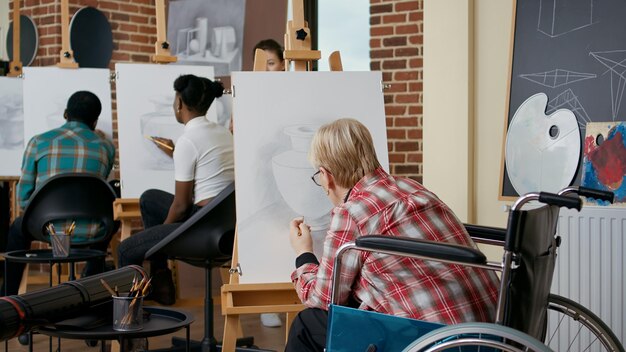Senior vrouw in rolstoel die vaasschets trekt met potlood op canvas en ezel, het bijwonen van een kunstlesprogramma met studenten en leraar. Persoon met een handicap die leert tekenen voor groei.