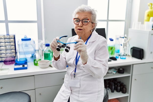 Gratis foto senior vrouw met grijs haar werken bij wetenschapper laboratorium met behulp van vergrootglazen puffende wangen met grappige gezicht mond opgeblazen met lucht vangen van lucht