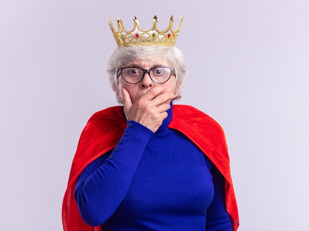 Gratis foto senior vrouw superheld dragen rode cape en bril met kroon op het hoofd kijken camera wordt geschokt die betrekking hebben op mond met hand permanent op witte achtergrond