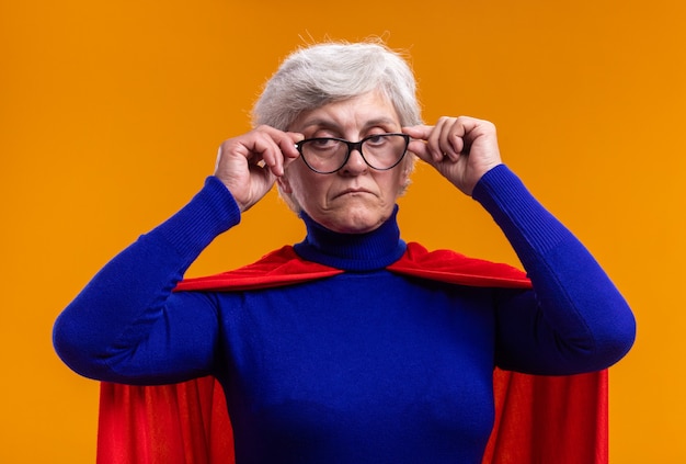 Gratis foto senior vrouw superheld met bril met rode cape kijken naar camera met ernstig gezicht staande over oranje achtergrond