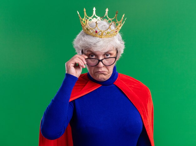 Senior vrouw superheld met rode cape en bril met kroon op het hoofd kijkend naar camera met sceptische uitdrukking staande over groene achtergrond