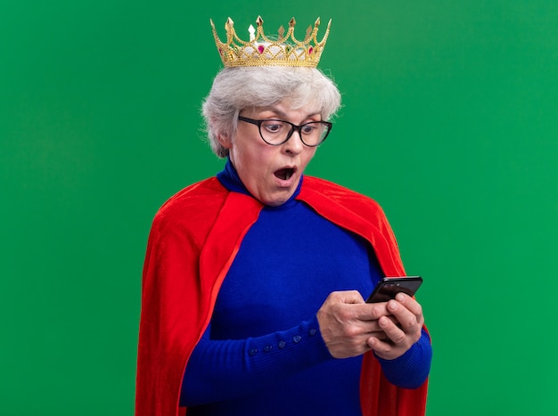 Senior vrouw superheld met rode cape en bril met kroon op het hoofd met behulp van smartphone verbaasd kijkend over groene achtergrond