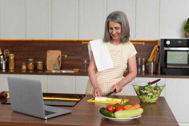 Senior vrouw thuis in de keuken die kooklessen volgt op laptop