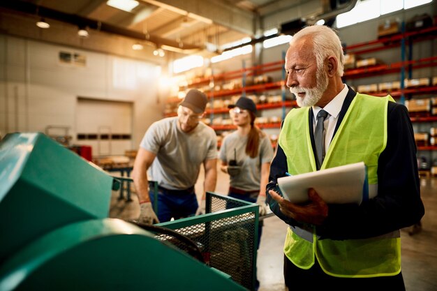 Senior zakenman die aantekeningen maakt tijdens het inspecteren van werknemers die aan de machine in het fabrieksmagazijn werken