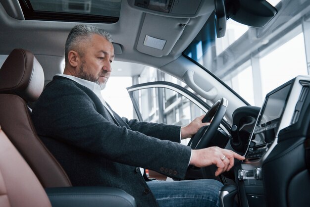 Senior zakenman in officiële kleding zit in een luxeauto en het indrukken van de knoppen op de muziekspeler