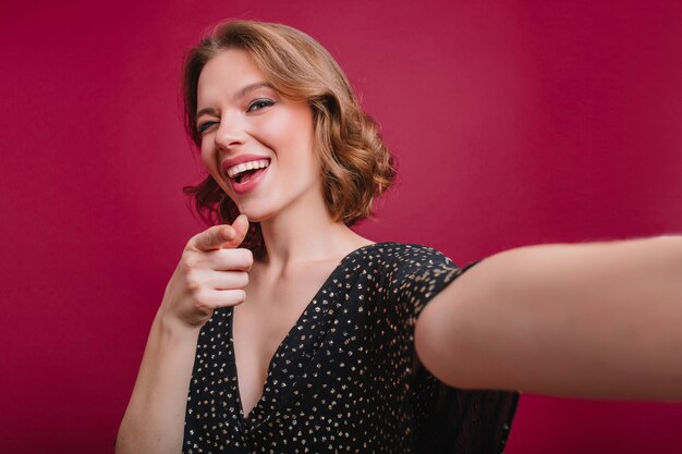 Sensueel meisje selfie maken Kortharige krullende vrouw in zwarte jurk nemen foto van zichzelf in kamer met paars interieur.