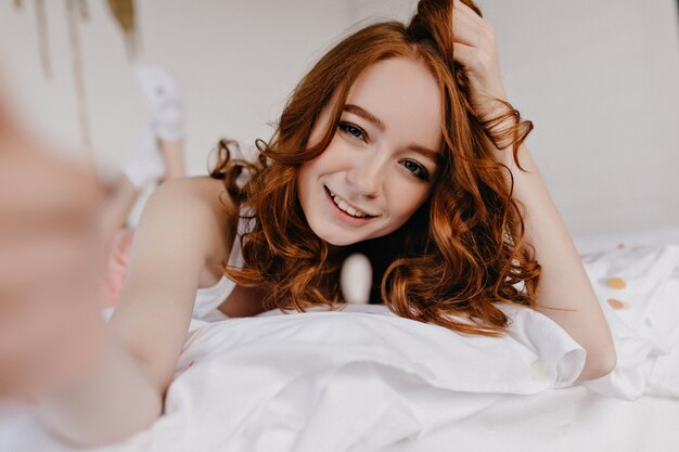 Sensueel wit meisje met golvend kapsel selfie maken in de ochtend. Aantrekkelijke jonge vrouw die op bed ligt en foto van zichzelf neemt.