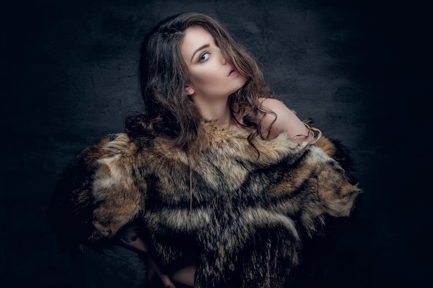 Sensuele brunette vrouw met krullend haar gekleed in een bontjas poseren in diepe schaduw op grijze achtergrond in een studio.