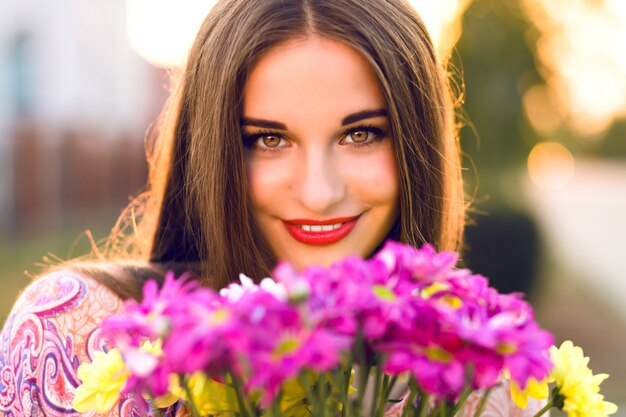 Sensuele brunette vrouw poseren met bloemboeket na romantische date, zonsondergang kleuren, elegante jurk en make-up.