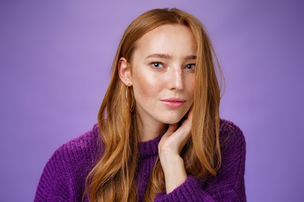 Sensuele en romantische vrouw met rood haar en sproeten die zachtjes de wang aanraken terwijl krullen op een half gezicht vallen en gecharmeerd en flirterig naar de camera staren, glimlachend zacht en koket over de paarse muur.