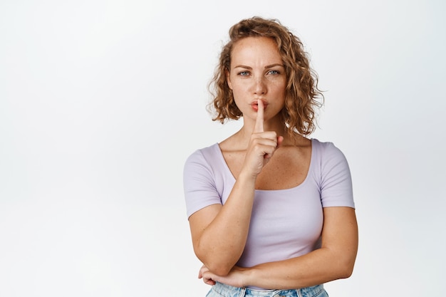 Serieus blond meisje dat zwijgt, zegt dat ze stil moet zijn, taboe shh-gebaar maakt met een boos gezicht op wit