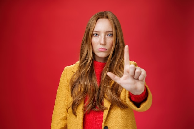 Serieus ogende roodharige vrouw in gele jas die hand naar camera uitstrekt met stop en waarschuwingsgebaar dat iets verbiedt en verbiedt, maakt iets met een emotieloos gezicht als zelfverzekerd over de rode muur.