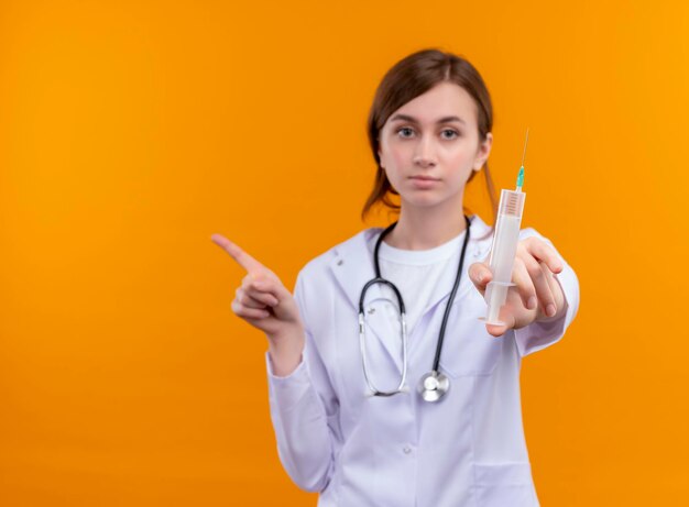 Serieus op zoek naar jonge vrouwelijke arts die medische mantel en stethoscoop draagt ?? die spuit uitrekt en naar de linkerkant wijst op geïsoleerde oranje ruimte met kopie ruimte