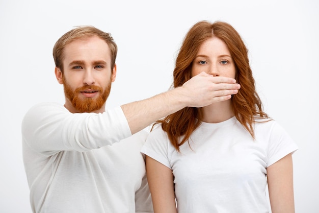 Gratis foto serieus roodharige man die over een witte muur staat en de mond van een roodharige vrouw verbergt