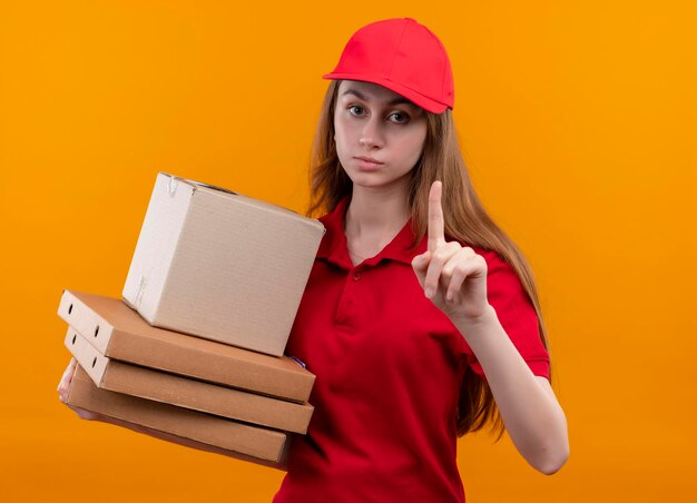 Serieus uitziend jong bezorgmeisje in rode uniforme holdingsdoos en pakketten met opgeheven vinger op geïsoleerde oranje ruimte