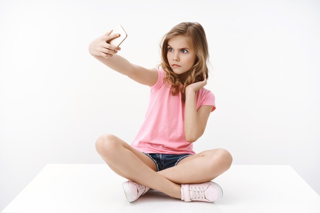 Serieus uitziende grappige schattige blonde Europese tienermeisje zittend op de vloer met gekruiste benen, strek arm smartphone uit om grimas te proberen, boos zelfverzekerde uitdrukking te maken, selfie te nemen, te fotograferen