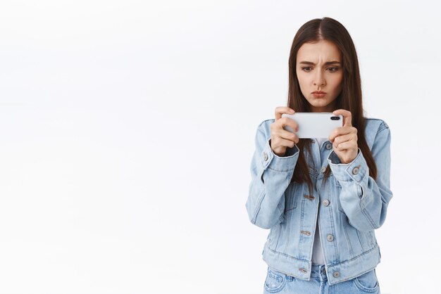 Serieus uitziende schattige en vrouwelijke blanke vrouw in denim met smartphone horizontaal pruilend vastberaden en gefocust als het spelen van een mobiel spel dat een moeilijk niveau witte achtergrond probeert te passeren