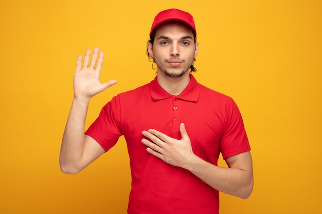 serieuze jonge bezorger met uniform en pet kijkend naar camera met belofte gebaar geïsoleerd op gele achtergrond