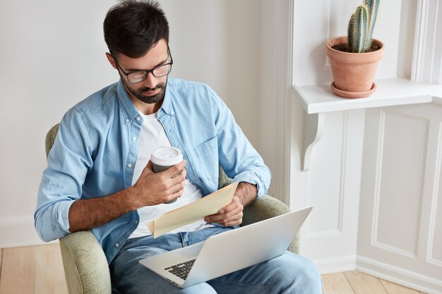 Serieuze ondernemer zorgt voor zaken, werkt thuis, concentreert zich op documenten, heeft laptop op knieën, houdt koffie voor afhaalmaaltijden, zit in comfortabele fauteuil.