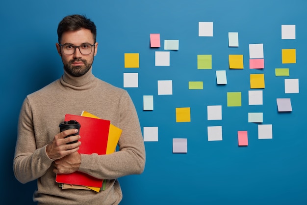 serieuze ongeschoren man drinkt afhaalkoffie, kleurrijke blanco stickers aan de muur, drinkt koffie uit papieren beker, houdt blocnotes vast