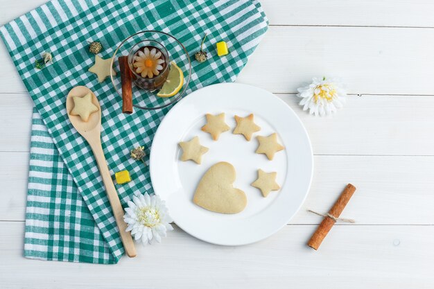 Set koekjes, citroen, kaneelstokje, suikerklontjes, bloemen en thee in een glas op houten en keukenhanddoekachtergrond. bovenaanzicht.