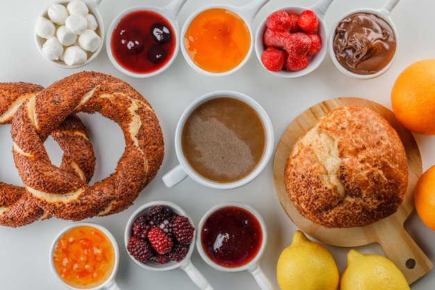 Set van jam, framboos, suiker, chocolade in kopjes, Turkse bagel, brood, sinaasappel en citroenen en een kopje koffie op een witte ondergrond