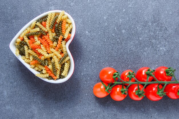 Set van tomaten en kleurrijke macaroni pasta in een hartvormige kom op een grijze ondergrond. bovenaanzicht. ruimte voor tekst