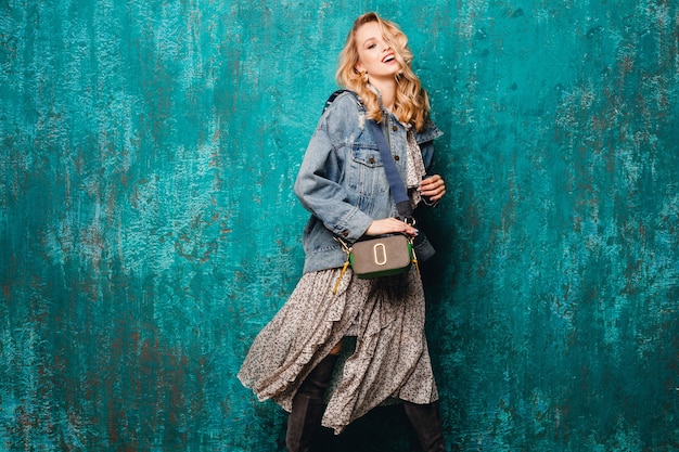 Sexy aantrekkelijke stijlvolle blonde vrouw in spijkerbroek en oversized jas lopen tegen vintage groene muur in straat