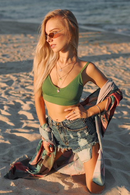 Sexy blonde vrouw in groene crop top en jeans poseren op tropisch strand.