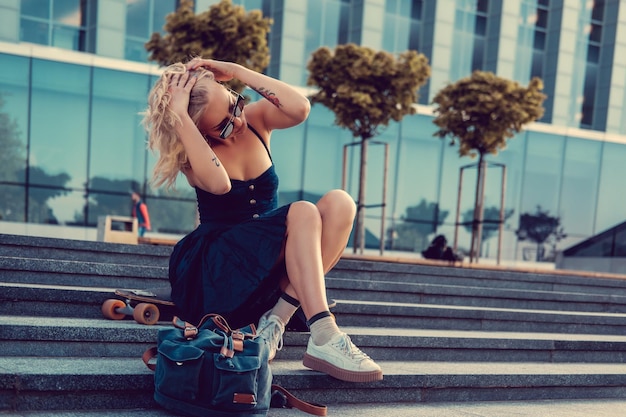 Gratis foto sexy blonde vrouw in zwarte jurk poseren met longboard op trappen in het centrum.