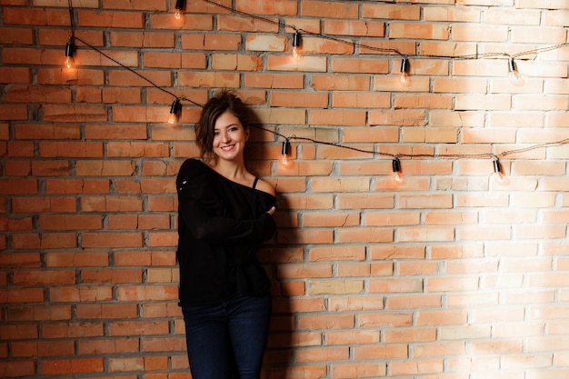 Sexy mooie vrouw in zwarte sweater en jeans die zich dichtbij een retro rode bakstenen muur bevinden