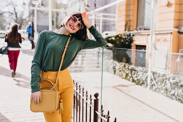 Shapely brunette vrouw met lichte make-up staande op straat. Outdoor Portret van gelukkig meisje met trendy kapsel poseren in lentestad.