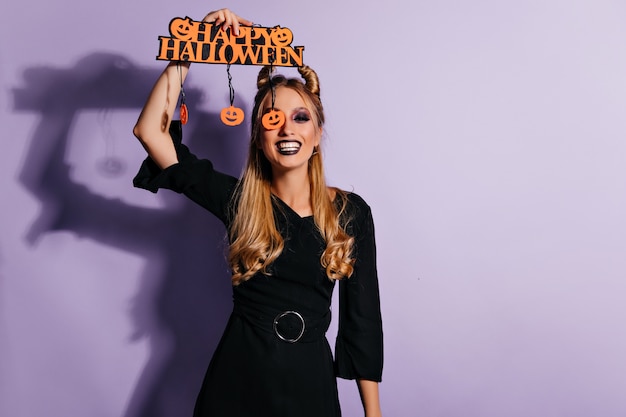 Shapely gelukkig meisje lachend in halloween. Indoor foto van prachtig wit vrouwelijk model in zwarte jurk geïsoleerd op pastel muur.