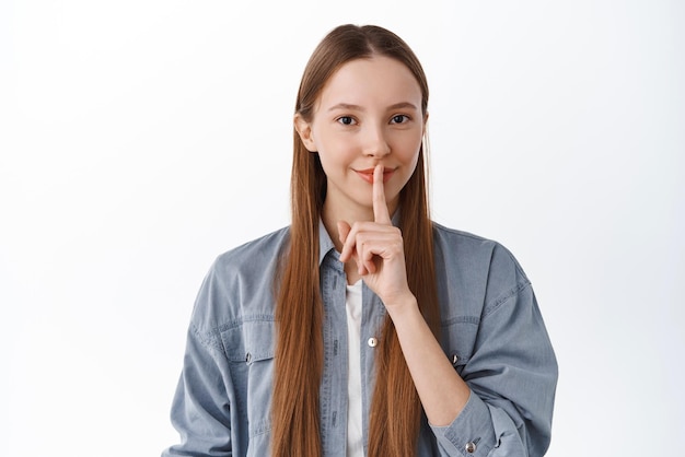 Gratis foto shh, dat is een geheim dat ik nooit vertel glimlachend schattig meisje dat iets verbergt en vraagt om stil te blijven en een stil gebaar maakt, een stil teken dat op een witte achtergrond staat