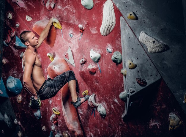 Shirtless atletische man klimmen op een rode indoor klimmuur.