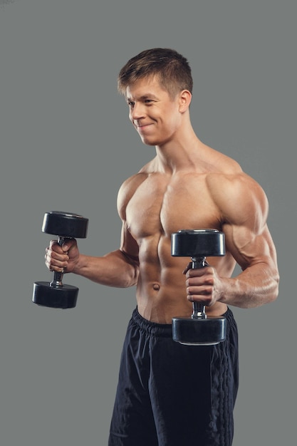 Shirtless gespierde man doet biceps-oefeningen met halters op een grijze achtergrond.