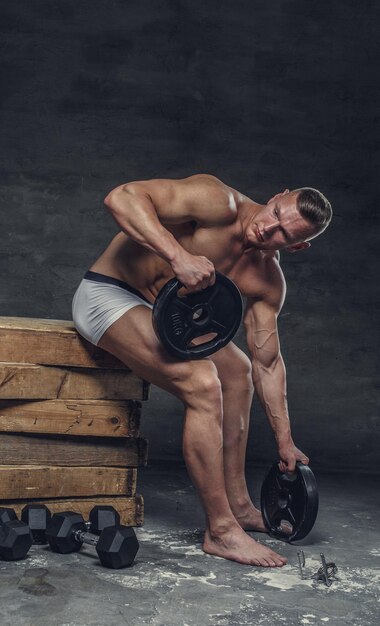 Shirtless gespierde man in een wit slipje zit op een houten kist met trainingsgewichten.