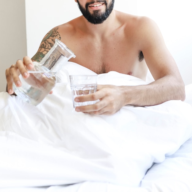 Gratis foto shirtless man zittend op bed met een glas water