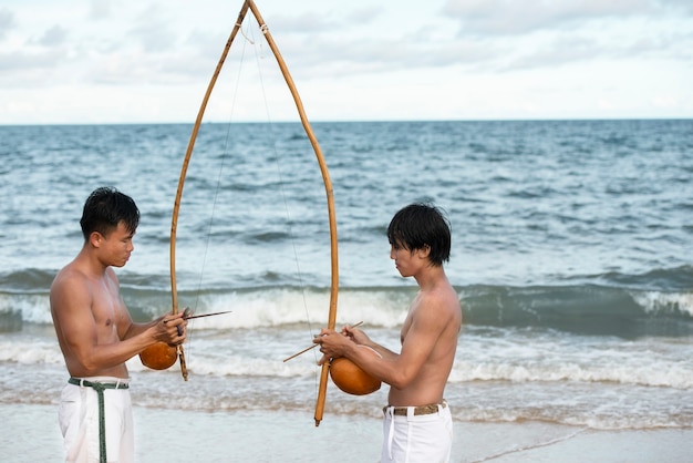 Shirtloze mannen die capoeira beoefenen op het strand met houten bogen