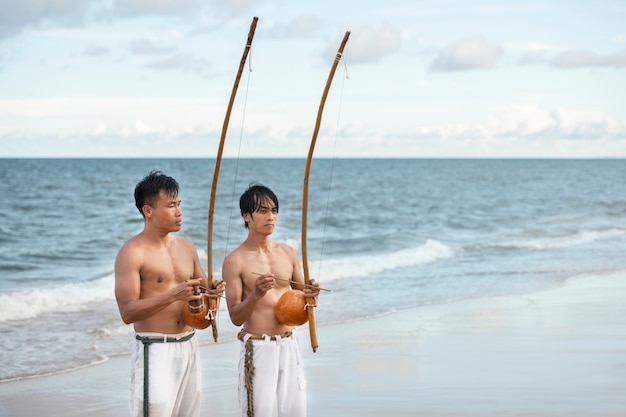 Gratis foto shirtloze mannen die capoeira beoefenen op het strand met houten bogen