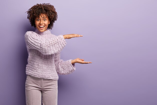 shot van blij Afro-Amerikaanse vrouw doet maat gebaar, toont iets groots, heeft krullend haar, gekleed in gebreide trui en broek, geïsoleerd op paarse muur. Bespreek kopie ruimte